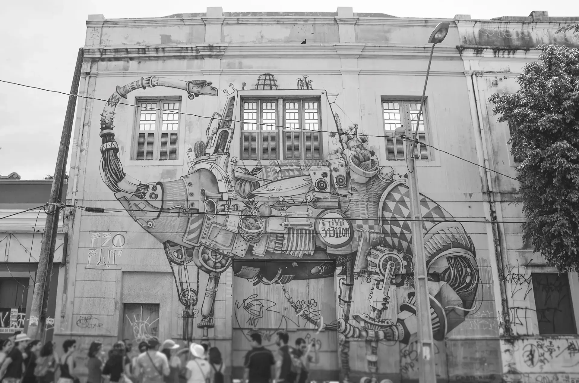 Fotografia em tons de cinza de um grande mural na fachada de um prédio antigo com um dinossauro futurista com vários acessórios sendo observado por um grupo de pessoas na frente do prédio