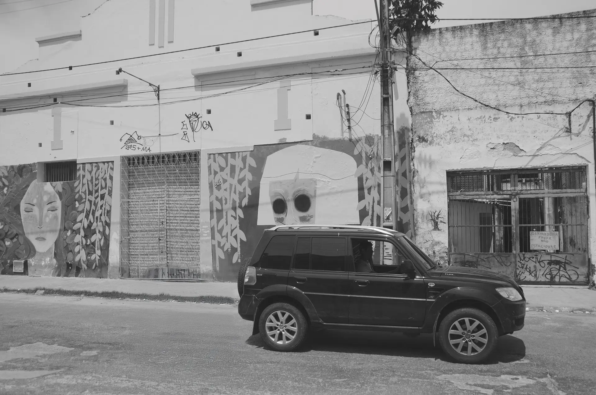 Frame 1 em tons de cinza de uma sequencia que mostra um carro atravessando uma rua no Centro de Fortaleza da esquerda para a direita com um mural de graffiti ao fundo