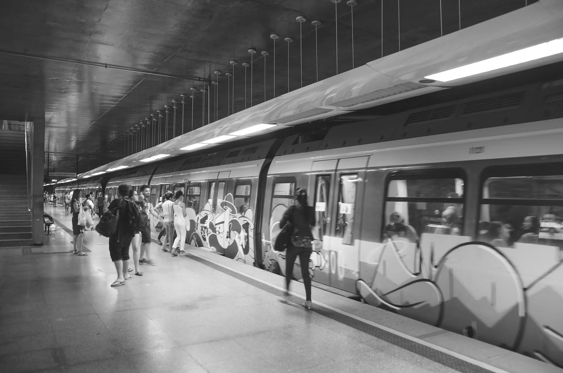 Fotografia em tons de cinza de uma mulher andando até a porta do vagão na Estação de metrô do Benfica em Fortaleza onde é possível observar os vagões grafitados do trem