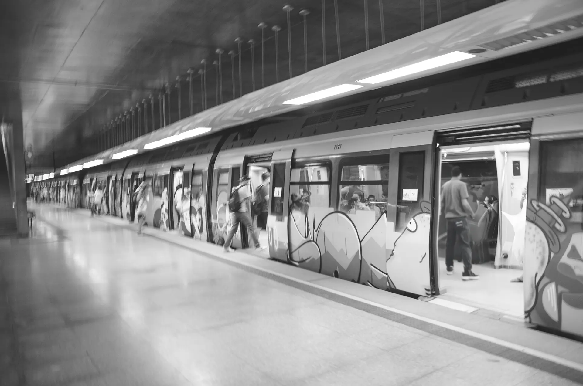 Fotografia em tons de cinza do trem grafitado e com as portas abertas na Estação de metrô do Benfica em Fortaleza enquanto pasageiros entram e saem do vagão