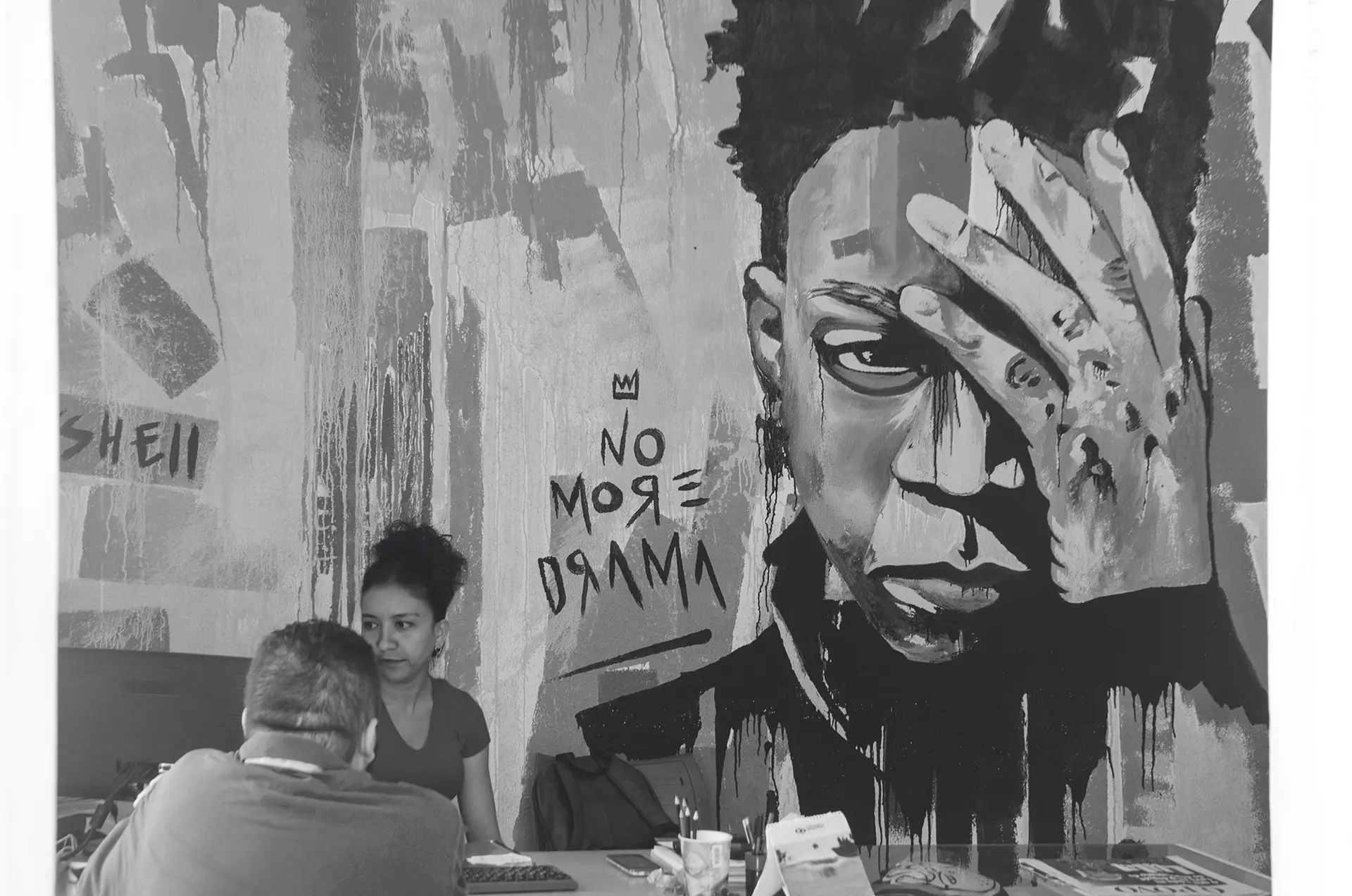 Fotografia em tons de cinza da recepção de uma escola de design com um mural de graffiti de Basquiat com a mão na testa e a frase NO MORE DRAMA enquanto uma recepcionista atende um aluno