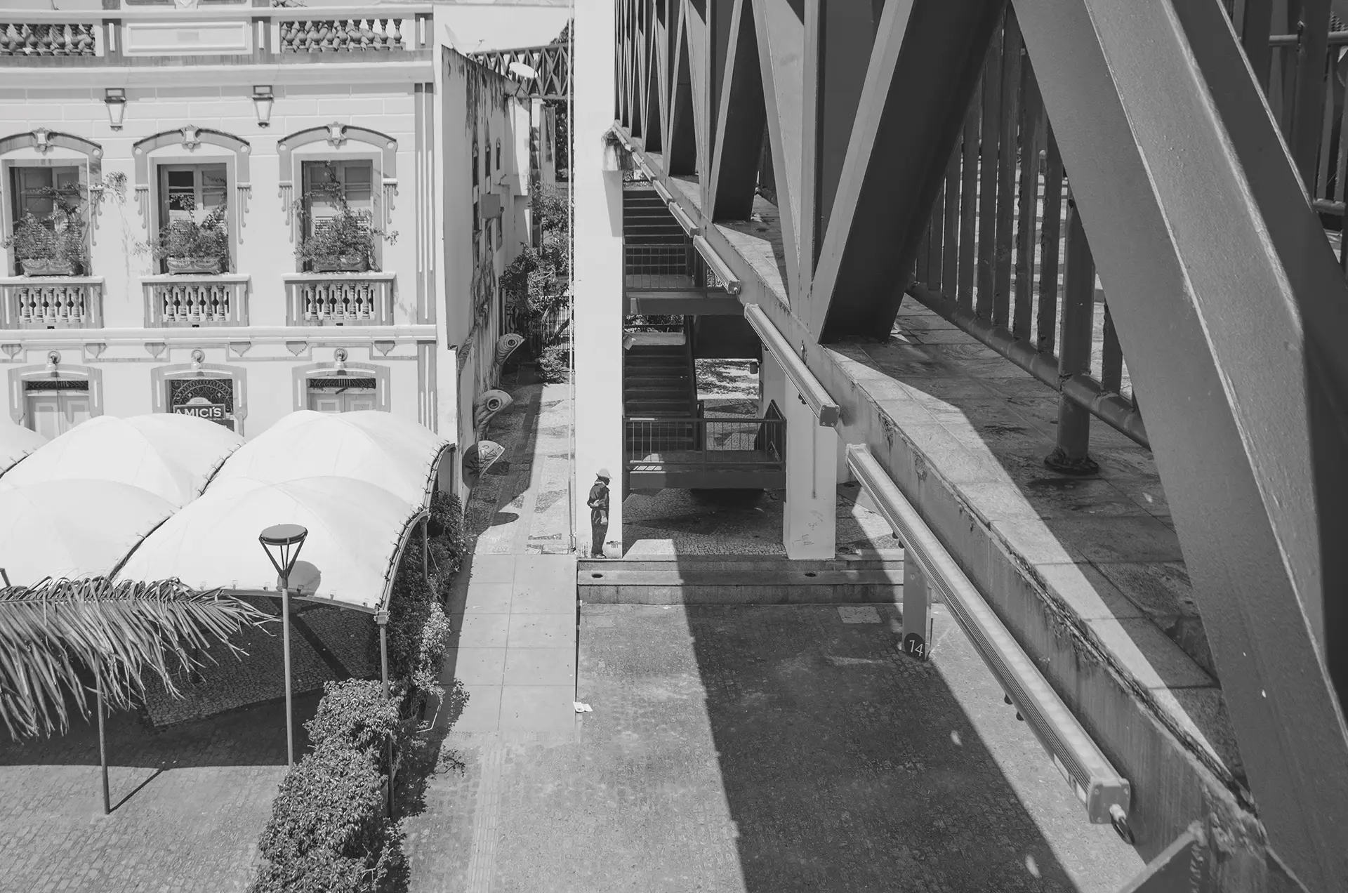 Fotografia em tons de cinza em perspectiva de uma das passarelas do Centro Cultural Dragão do Mar de Arte e Cultura em Fortaleza onde é possível observar a silhueta de um homem de costa e com os braços cruzados para atrás que foi feito com stencil na coluna da passarela