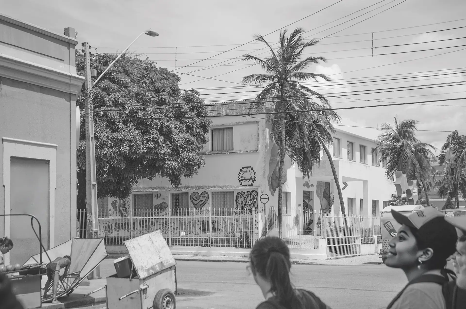Fotografia em tons de cinza mais distante de um prédio público com várias intervenções de arte urbana com uma mulher de costa observando o prédio