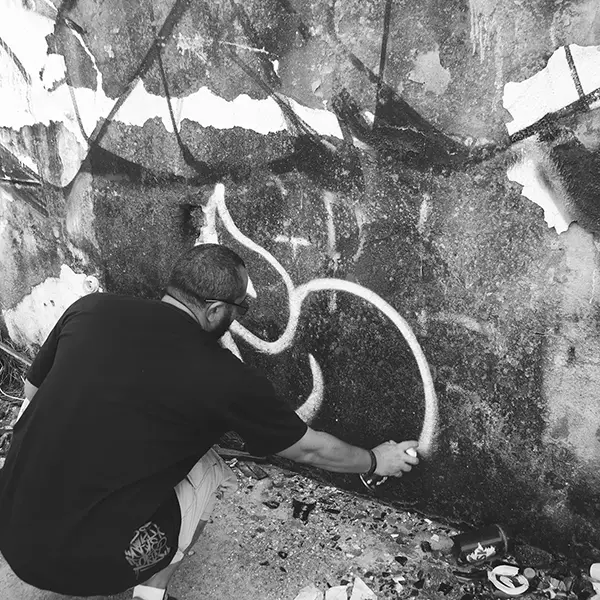 Frame 11 em tons de cinza de um vídeo de um homem de cócoras com uma lata de spray na mão enquanto desenha uma assinatura em um muro de um prédio abandonado