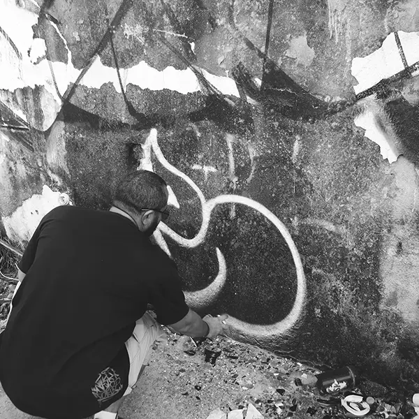 Frame 12 em tons de cinza de um vídeo de um homem de cócoras com uma lata de spray na mão enquanto desenha uma assinatura em um muro de um prédio abandonado