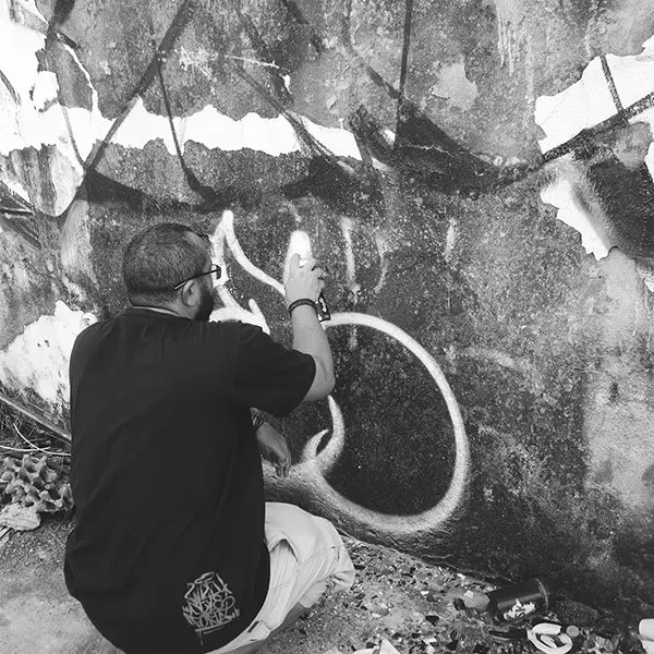 Frame 14 em tons de cinza de um vídeo de um homem de cócoras com uma lata de spray na mão enquanto desenha uma assinatura em um muro de um prédio abandonado