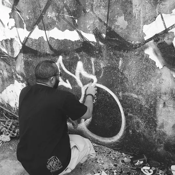 Frame 15 em tons de cinza de um vídeo de um homem de cócoras com uma lata de spray na mão enquanto desenha uma assinatura em um muro de um prédio abandonado