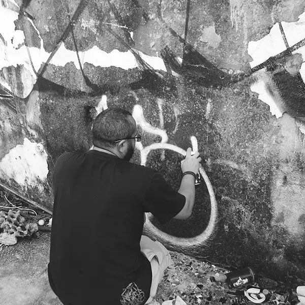 Frame 16 em tons de cinza de um vídeo de um homem de cócoras com uma lata de spray na mão enquanto desenha uma assinatura em um muro de um prédio abandonado