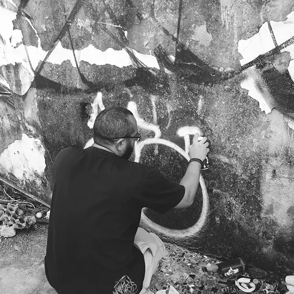 Frame 17 em tons de cinza de um vídeo de um homem de cócoras com uma lata de spray na mão enquanto desenha uma assinatura em um muro de um prédio abandonado