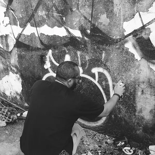 Frame 18 em tons de cinza de um vídeo de um homem de cócoras com uma lata de spray na mão enquanto desenha uma assinatura em um muro de um prédio abandonado