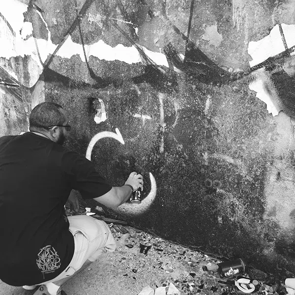 Frame 2 em tons de cinza de um vídeo de um homem de cócoras com uma lata de spray na mão enquanto desenha uma assinatura em um muro de um prédio abandonado