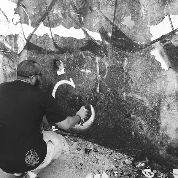 Frame 3 em tons de cinza de um vídeo de um homem de cócoras com uma lata de spray na mão enquanto desenha uma assinatura em um muro de um prédio abandonado