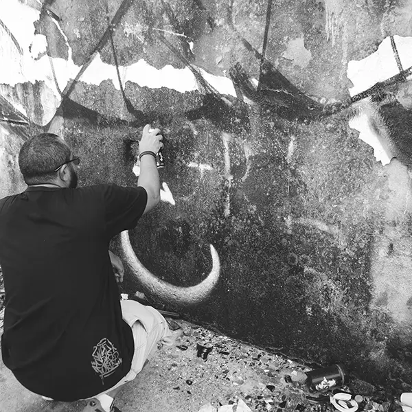 Frame 5 em tons de cinza de um vídeo de um homem de cócoras com uma lata de spray na mão enquanto desenha uma assinatura em um muro de um prédio abandonado
