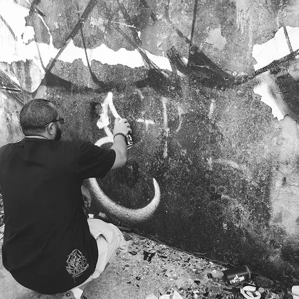 Frame 6 em tons de cinza de um vídeo de um homem de cócoras com uma lata de spray na mão enquanto desenha uma assinatura em um muro de um prédio abandonado