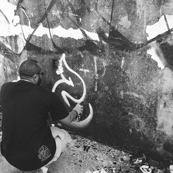 Frame 9 em tons de cinza de um vídeo de um homem de cócoras com uma lata de spray na mão enquanto desenha uma assinatura em um muro de um prédio abandonado